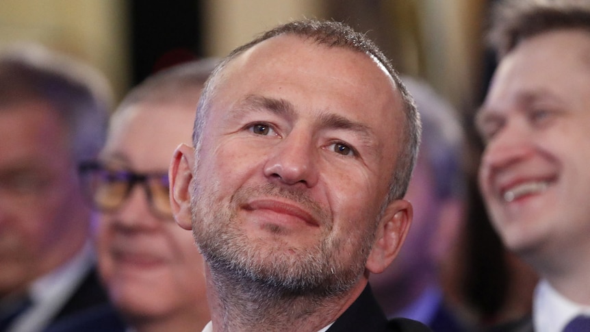 A middle-aged, short-haired white man with stubble in a suit looks forward while seated with a group of people.