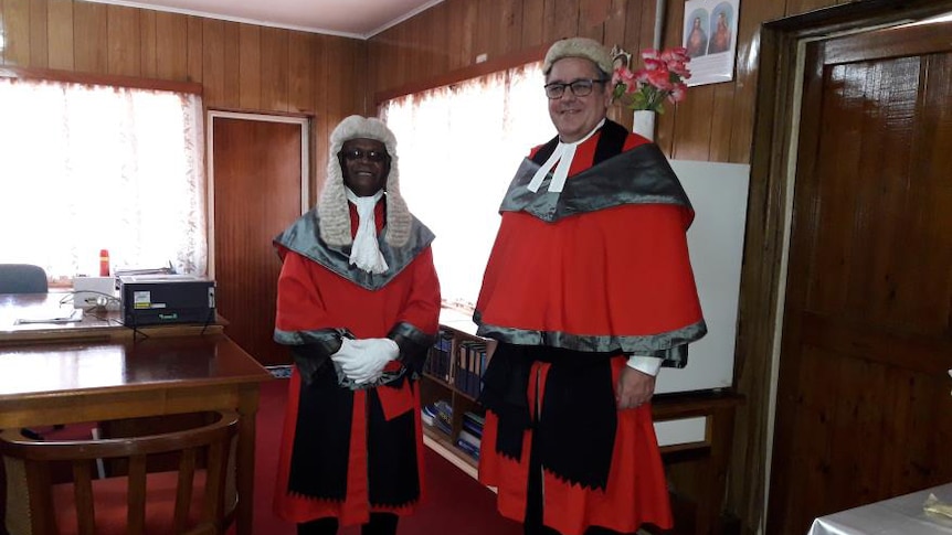Two men stand in high court judge gowns and wigs. 