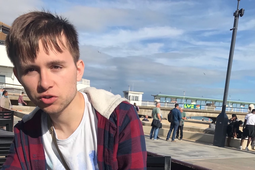 A young man with brown hair sits on a foreshore 