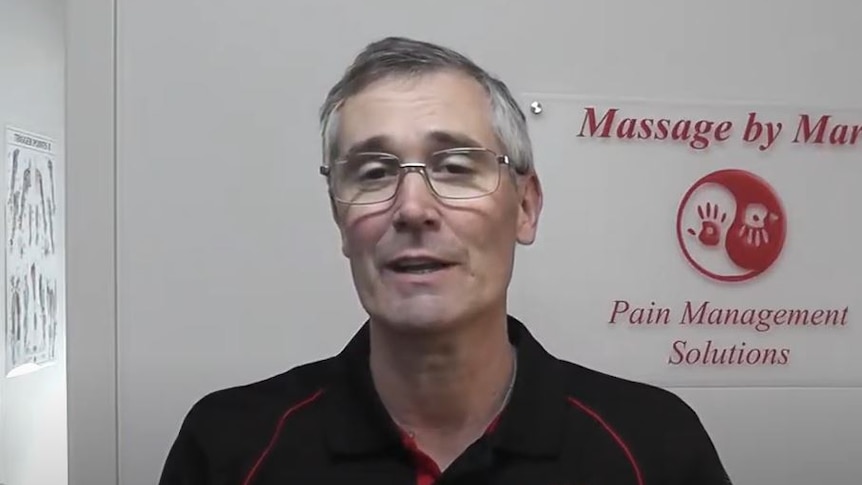A man with grey hair and glasses wearing a black polo shirt with a logo on the wall behind him