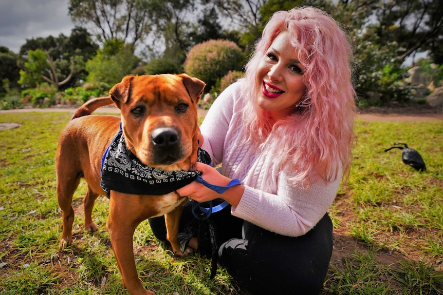 A woman sits on the grass, smiling at the camera and holding her rescue dog