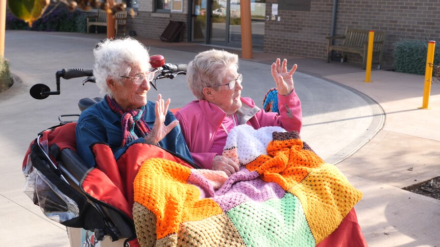 Two elderly women sitting in the front of a trishaw waving with a coloured blanket on their lap.