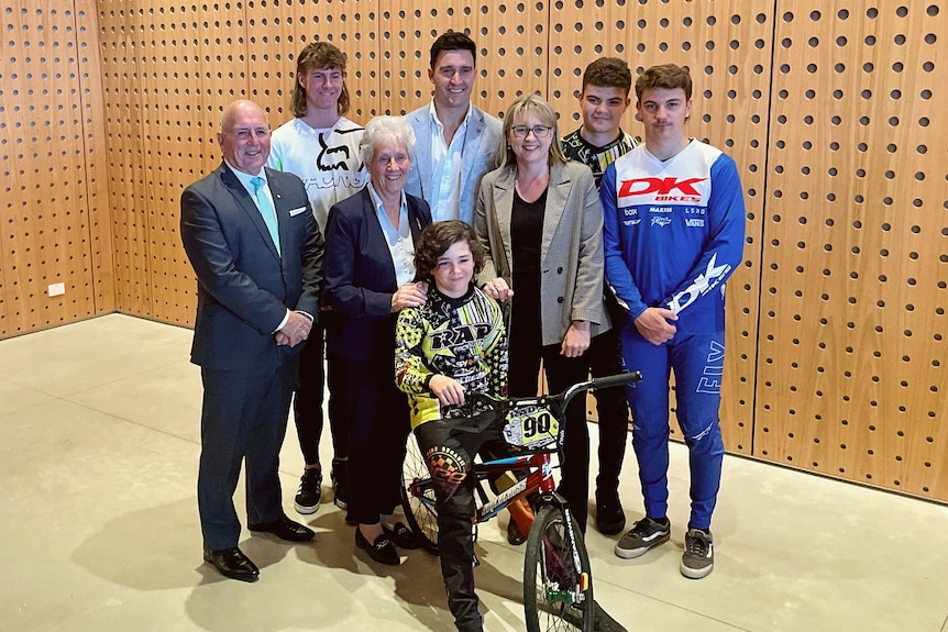 A group of smiling adults stand around a child on a BMX.