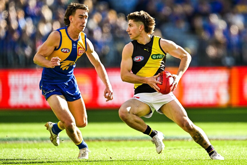 A Richmond AFL player holds the ball in two hands as he attempts to evade a West Coast opponent.
