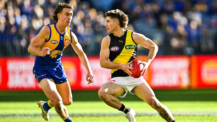 A Richmond AFL player holds the ball in two hands as he attempts to evade a West Coast opponent.