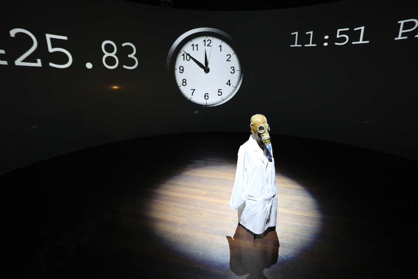 A figure wearing a gas mask and lab coat stands in a pool of light, in front of a clock set at nine minutes to midnight.