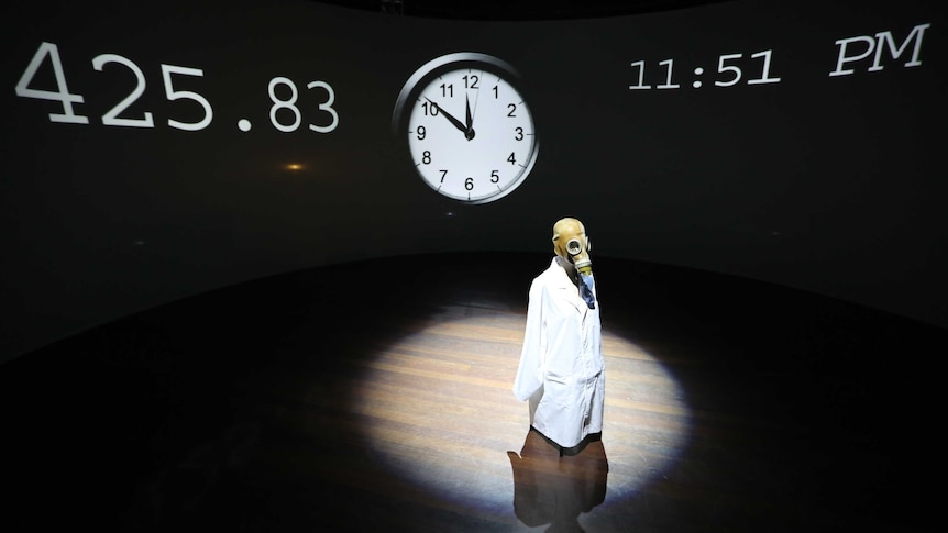 A figure wearing a gas mask and lab coat stands in a pool of light, in front of a clock set at nine minutes to midnight.