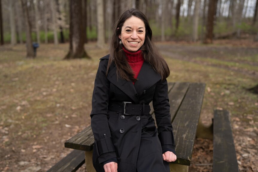A woman with long hair in a red turtleneck and coat smiles at camera while leaning against a park bench