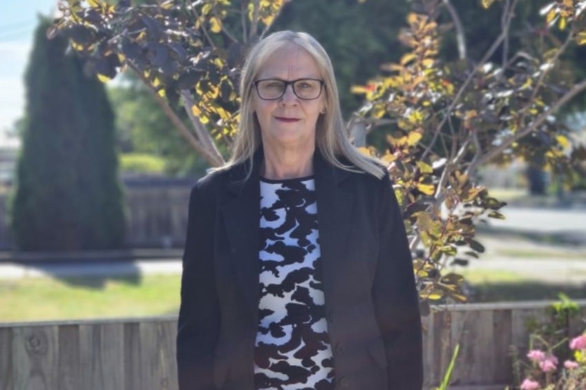 A serious woman with blonde hair, glasses, black jacket, black and white top stands in front of a green bush.