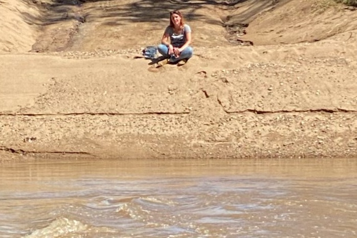 A woman sits cross legged on a pile of sand with floodwater covering the submerged bridge in front of her.