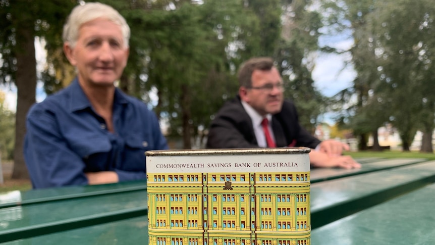 Two men sit at a table, with a Commonwealth Bank tin money box in the foreground.