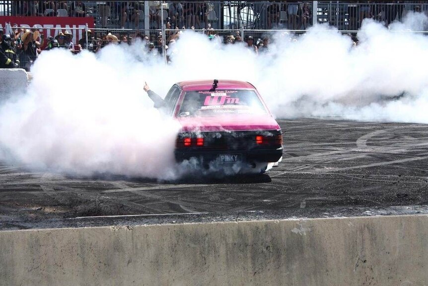 Rear view of a pink 1986 Holden Commodore with a PINKY number plate doing a burnout while a hand is sticking out the window 