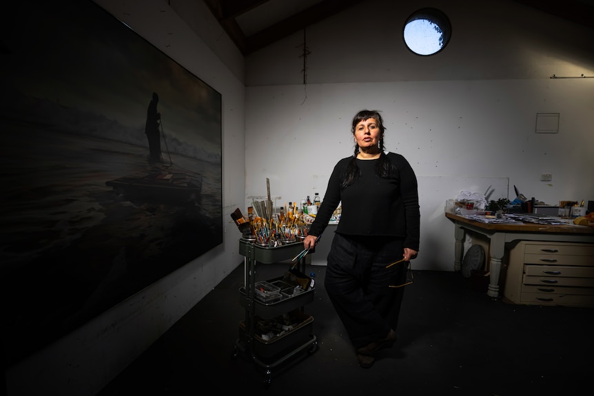 50-something artist dressed in black with long dark hair in plaits stands in darkened studio with paint brushes to hand.