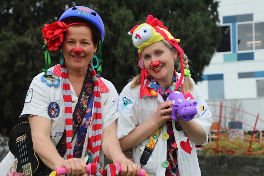 Two women dressed in clown costumes smile, one holding a toy cow and the other a guitar.