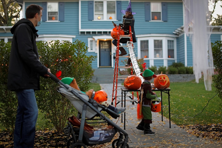 A woman sits on a ladder dressed as a witch and drops candy down a tube to a little boy waiting outside her gate.