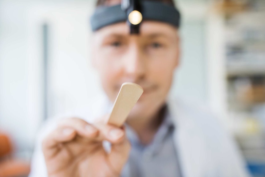 A tongue depressor in the foreground, with a doctor wearing a headlamp out of focus in the background.