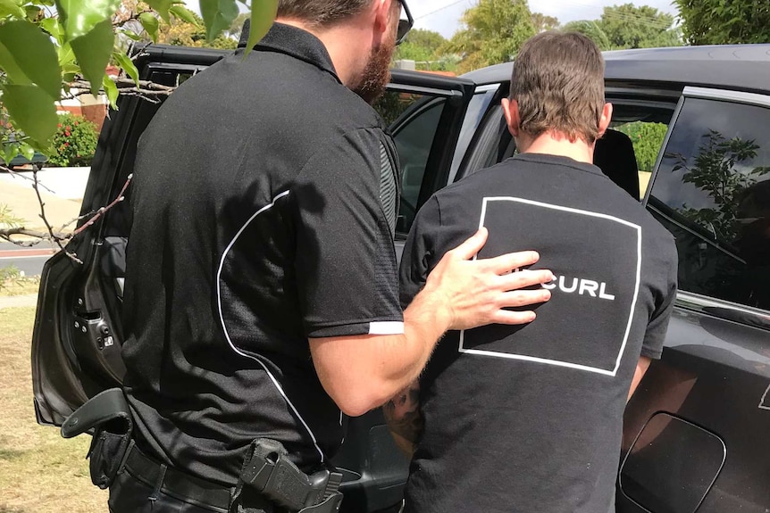 A man in a black shirt is escorted into a waiting car by a plain-clothes police officer.