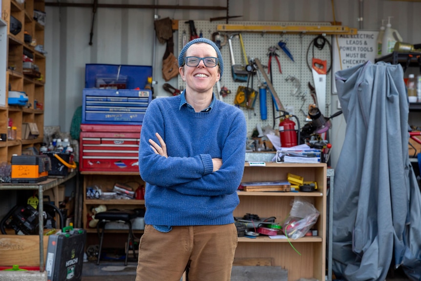 A woman with glasses, a blue jumper and brown trousers stands smiling with folded arms in front of a tool shed.