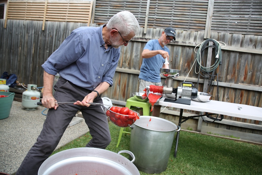 a man shovels tomatoes into a pot with a large strainer while another feeds them through a juicer
