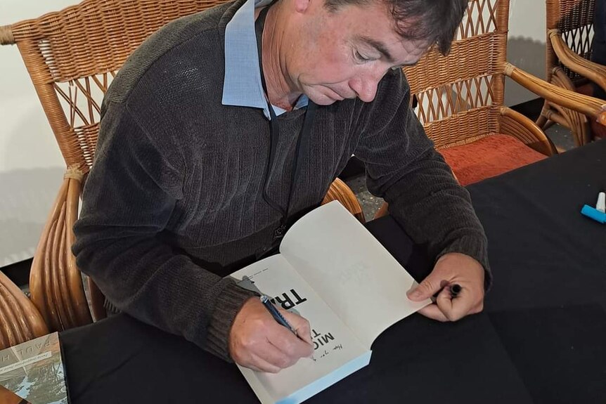 Overhead shot of a middle-aged man with dark hair, on a rattan chair, signs a book on cloth-covered table. Wears brown jumper.