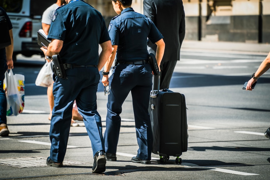 Two armed police officers in uniform cross a CBD street, one wheeling a suitcase while the other holds a folder in one arm.
