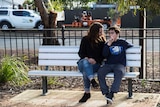 A woman and 14 year old boy sit on a bench, the boy is using sign to communicate.