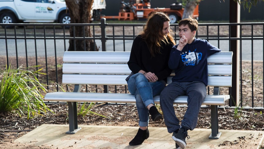 A woman and 14 year old boy sit on a bench, the boy is using sign to communicate.