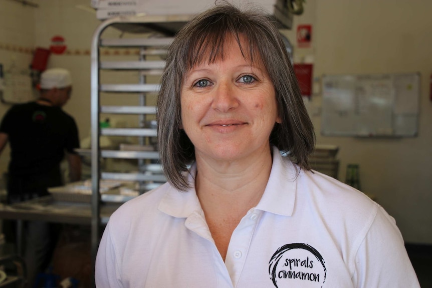 A middle-aged woman with greying hair stands in a cafe kitchen smiling for a photo wearing a white polo shirt.