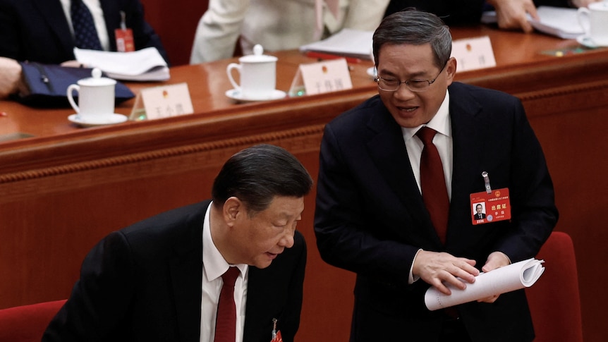 Li Qiang stands while holding a rolled up bunch of papers, next to Xi Jinping who is sitting