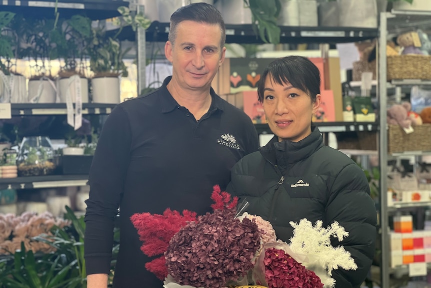 A man wearing a long sleeve collared black polo and a woman wearing a black puffer jacket stand in a florist.