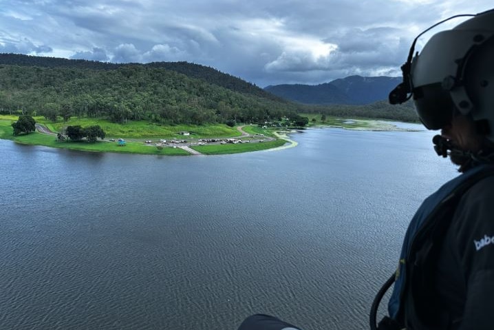 A CQ Rescue staff member sits at the side of a helicopter as it flies over a body of water.