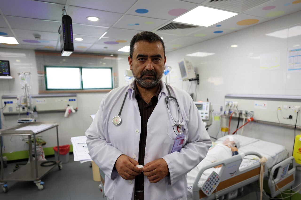 A medical professional stands inside a hospital and looks at the camera with a serious expression.