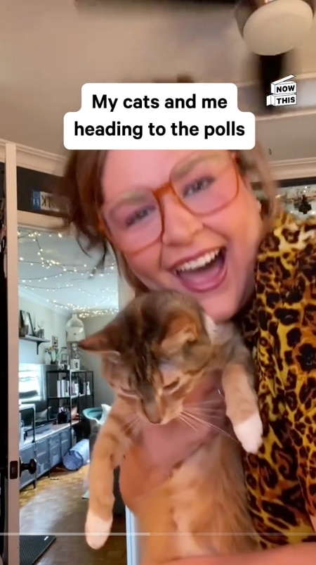 A still of a video where a woman in red glasses wearing a leopard print shirt holds up her cat and smiles.