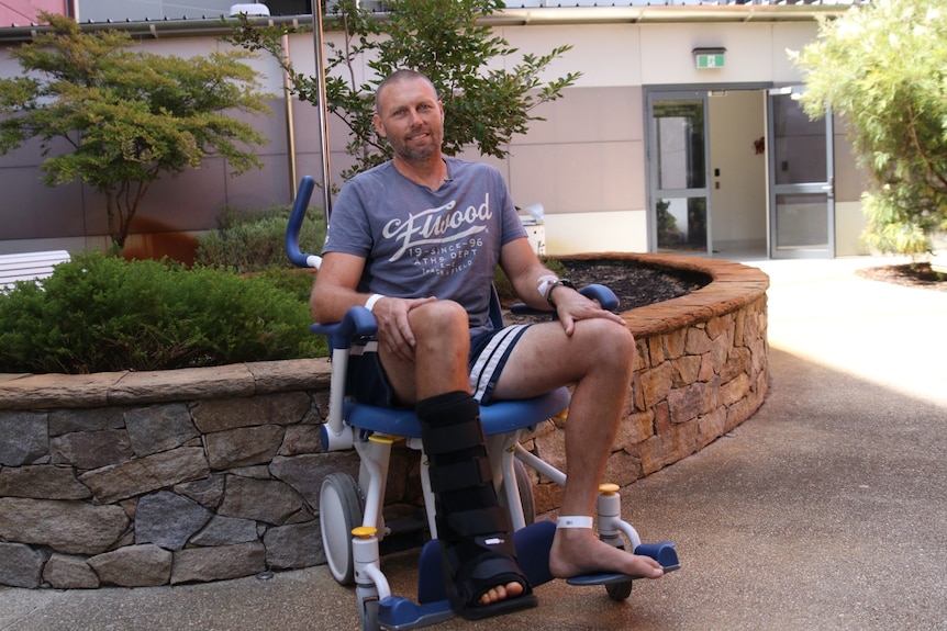 A smiling man sits in a wheelchair with a black moon boot on his right foot outside hospital.