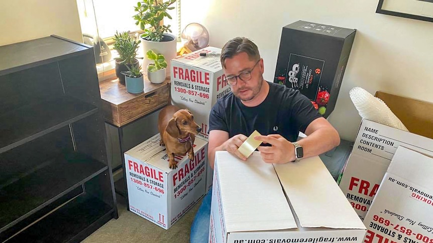 Paul Menz packing boxes, with his dog standing on one of the boxes.