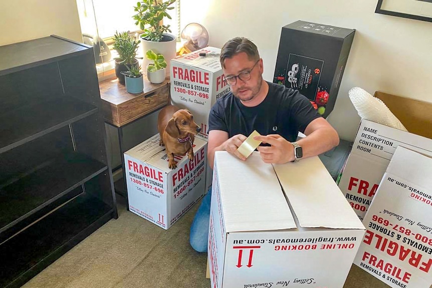Paul Menz packing boxes, with his dog standing on one of the boxes.
