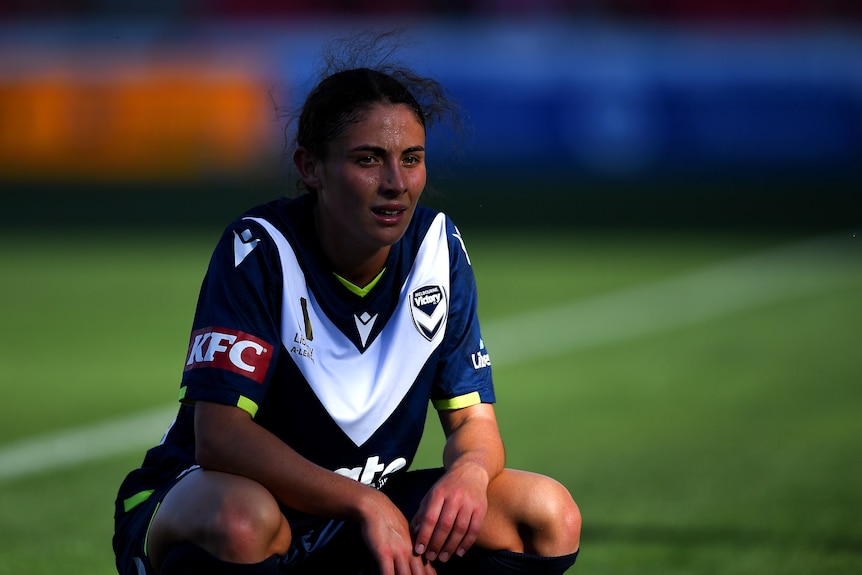 A soccer player wearing dark blue sits down and looks into the distance