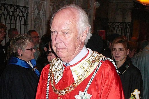 A man dressed in red robes with his family crest embroied on it stares off camera inside a cathedral.