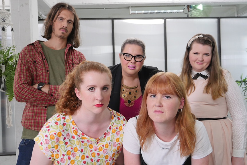 A group of four women and one man in an office space, display unimpressed expressions and look towards camera.