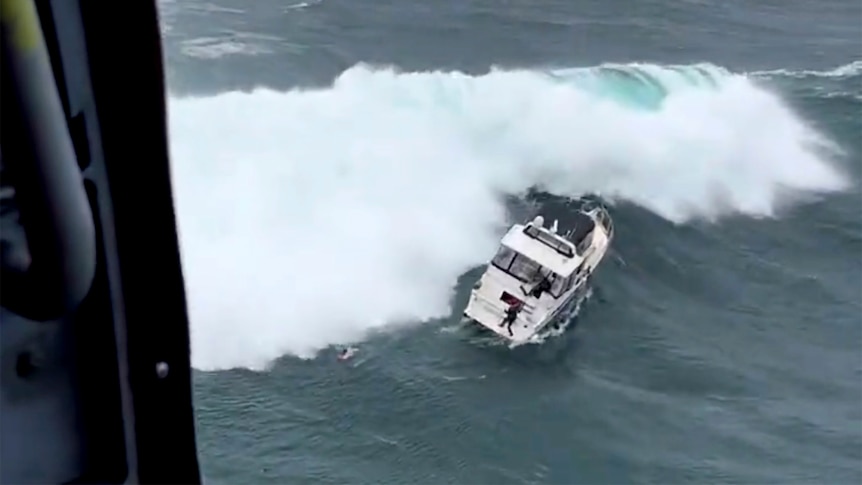 A massive wave bears down on a motor yacht. A man stands on the stern and a rescue swimmer is visible in the ocean.