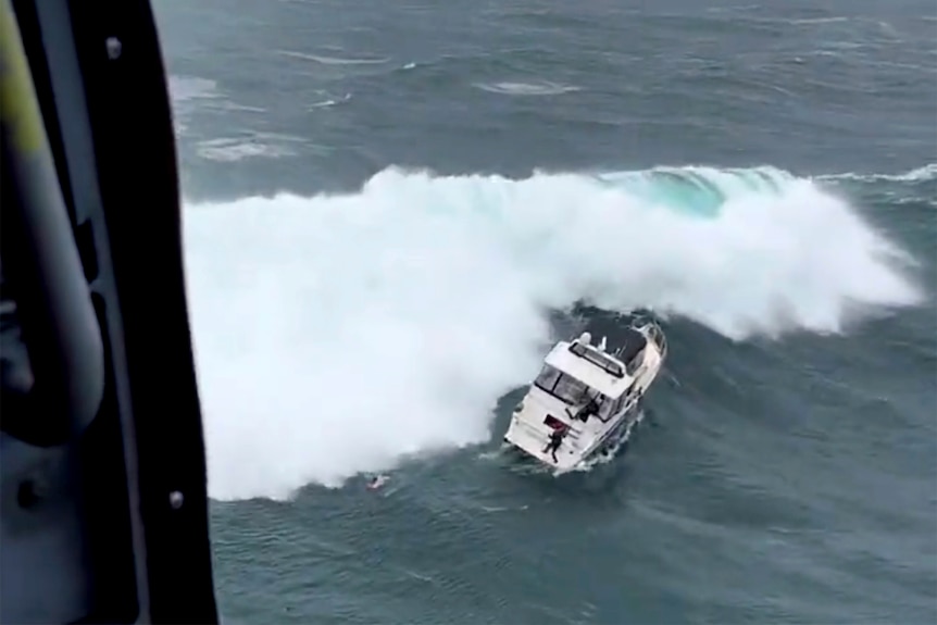 A massive wave bears down on a motor yacht. A man stands on the stern and a rescue swimmer is visible in the ocean.