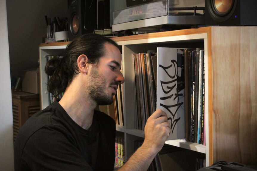 A man with a black shirt on sits in front of his record collection, pulling out a vinyl to look at
