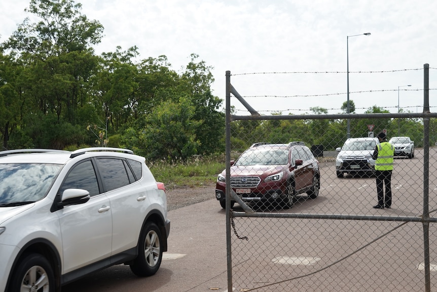 Cars line up to leave through a gate at Howard Springs facility. Someone is checking a clipboard as they drive past.