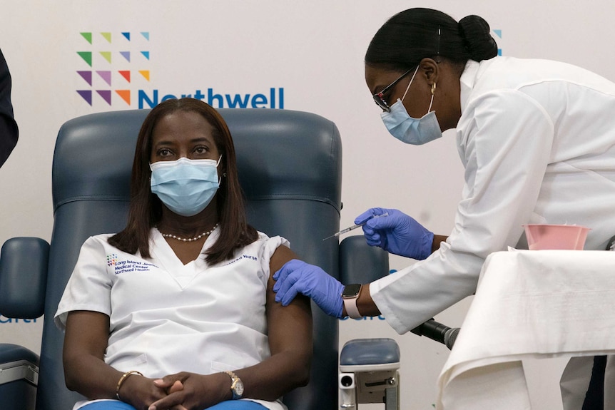 A black woman in a face mask sits while a black doctor with black hair tied in a bun injects her.
