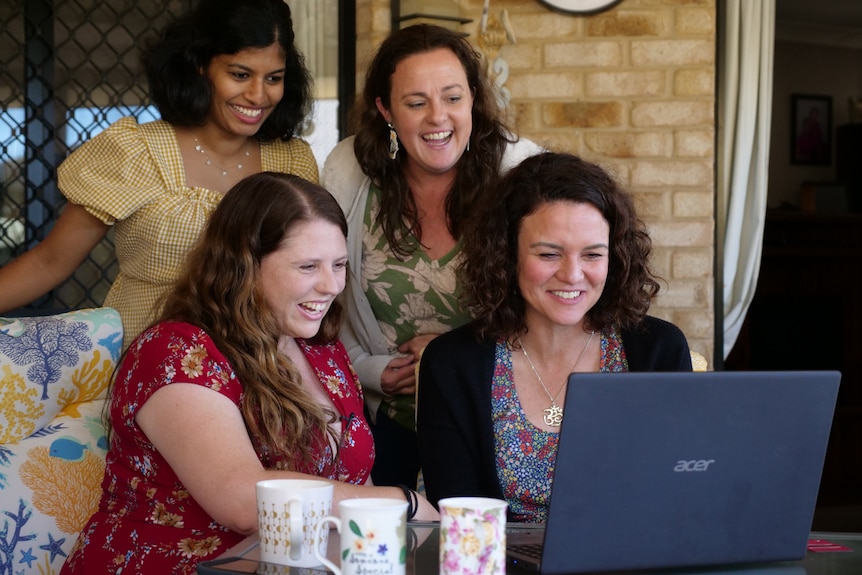 Angela, Simone, Saivashini and Bronwyn smiling at a laptop, mugs in the foreground.