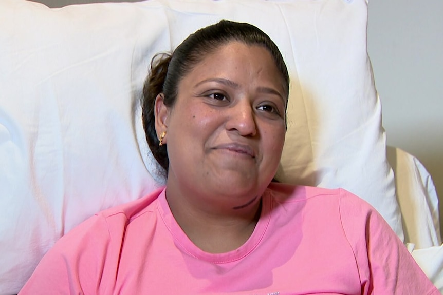 Roshni wearing a pink shirt laying in a hospital bed smiling.