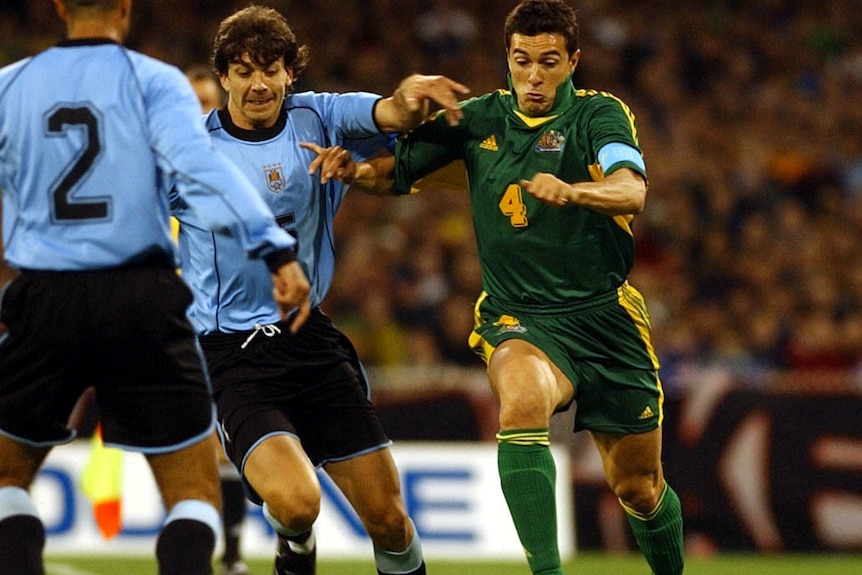 A Socceroos midfielder goes full tilt at the ball while a Uruguayan defender tries to pull him back.