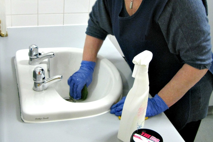 A cleaner works in a bathroom, cleaning a sink.