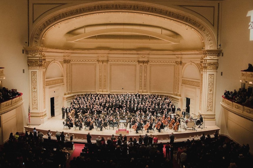 A wide shot of the choir and orchestra on stage at Carnegie hall, while in the foreground the audience gives a standing ovation.