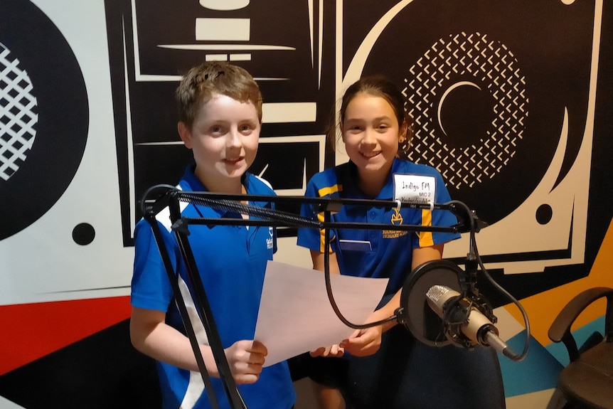 Flynn Graham and Emma Pinard stand behind a radio mic in a radio studio.